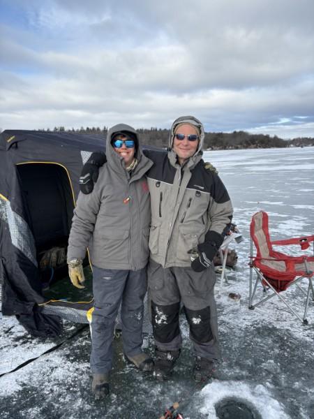 Two friends in winter gear standing by an ice fishing shanty